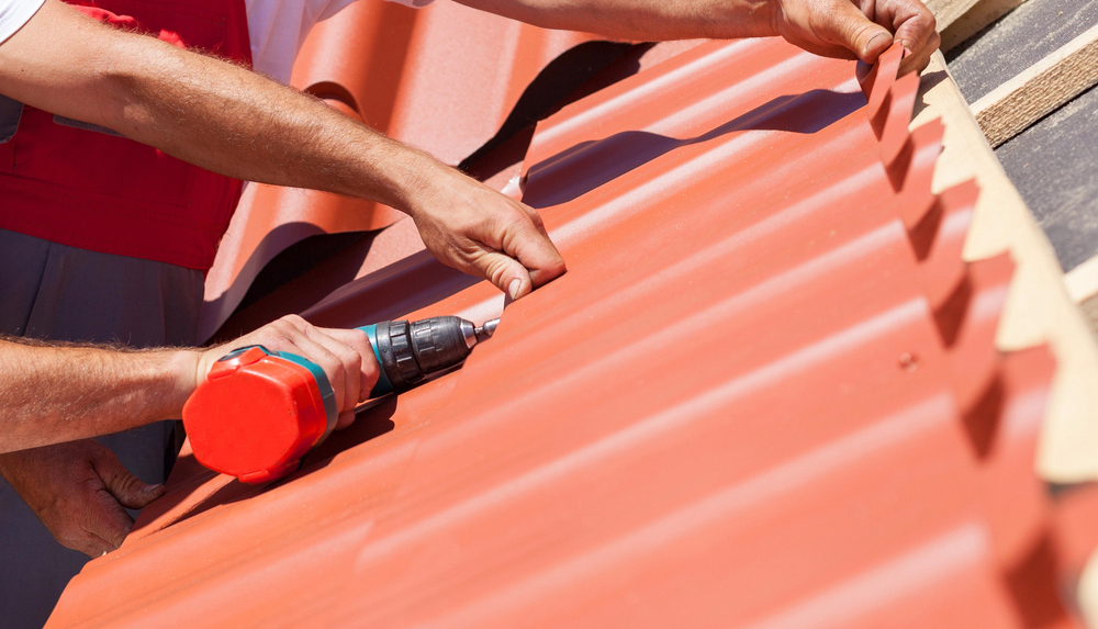 Workers on a roof with electric drill installing red metal tile 